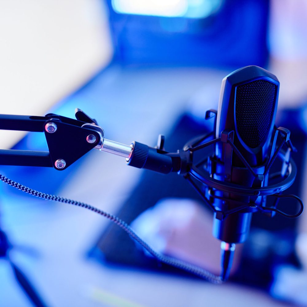 Close up of professional microphone on stand in video game streaming setup lit by blue neon light, copy space